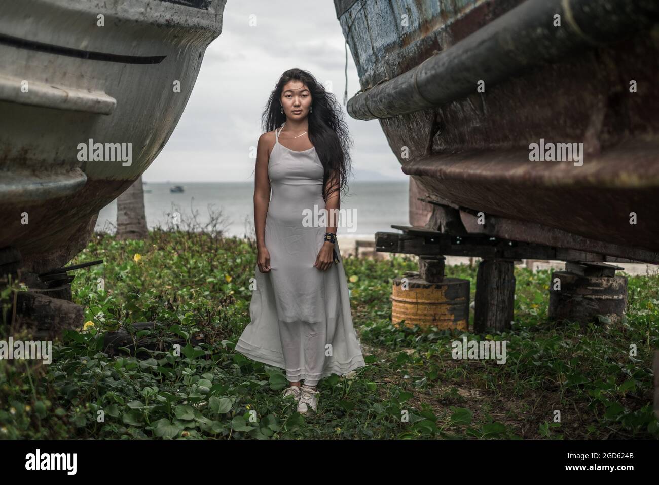 Charming young mongol woman in bright grey dress standing between two old wooden ships. Black long curly hair. Looking at camera with copy space. Stock Photo