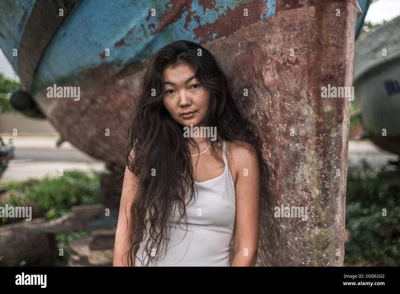 Portrait of beautiful young asian woman in white dress standing near the old ship. Natural black curly long hair. Thinking romantic face.  Stock Photo