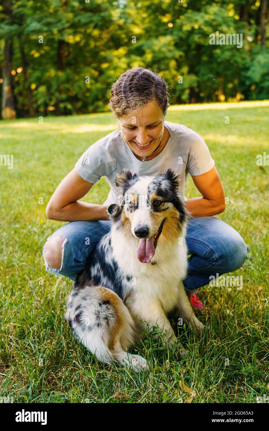 Happy young woman spending time with lovely spotted Australian Shepherd dog in green meadow on summer day Stock Photo