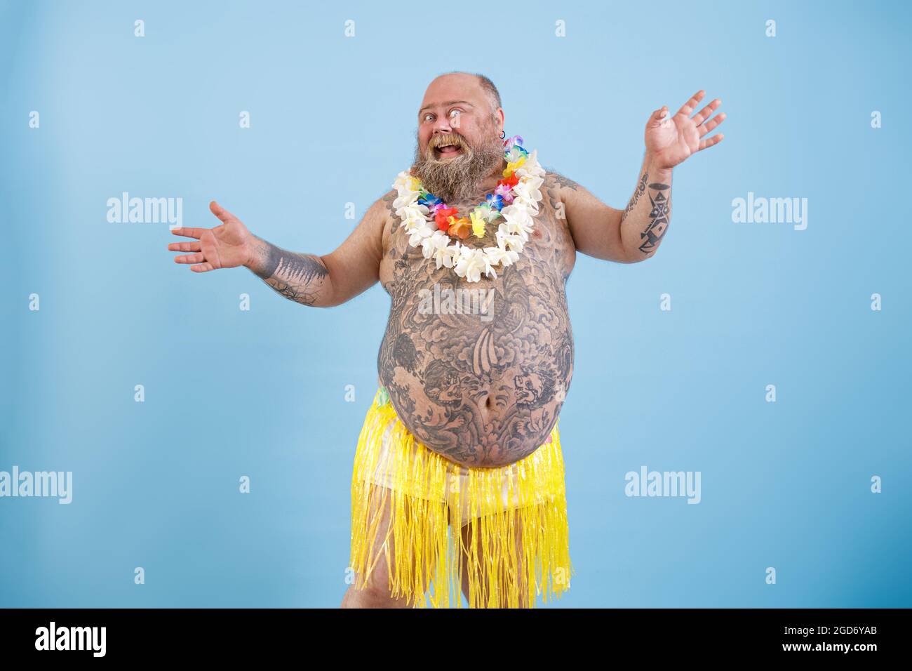 Positive fat man with tattoos in decorative grass skirt dances on light blue background Stock Photo
