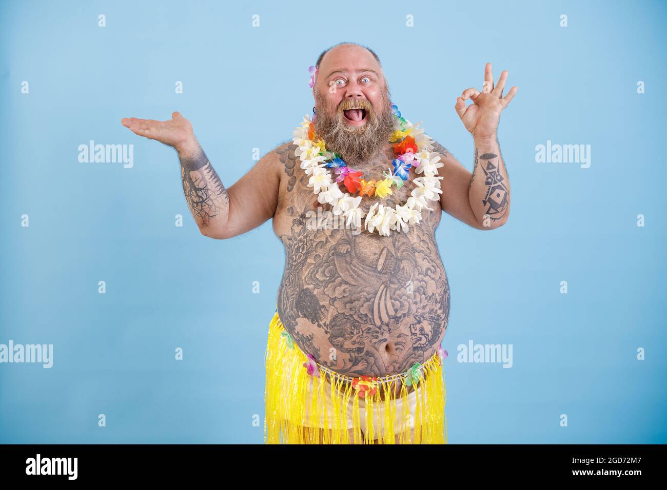Excited fat man in grass skirt points aside and shows Ok gesture on blue background Stock Photo