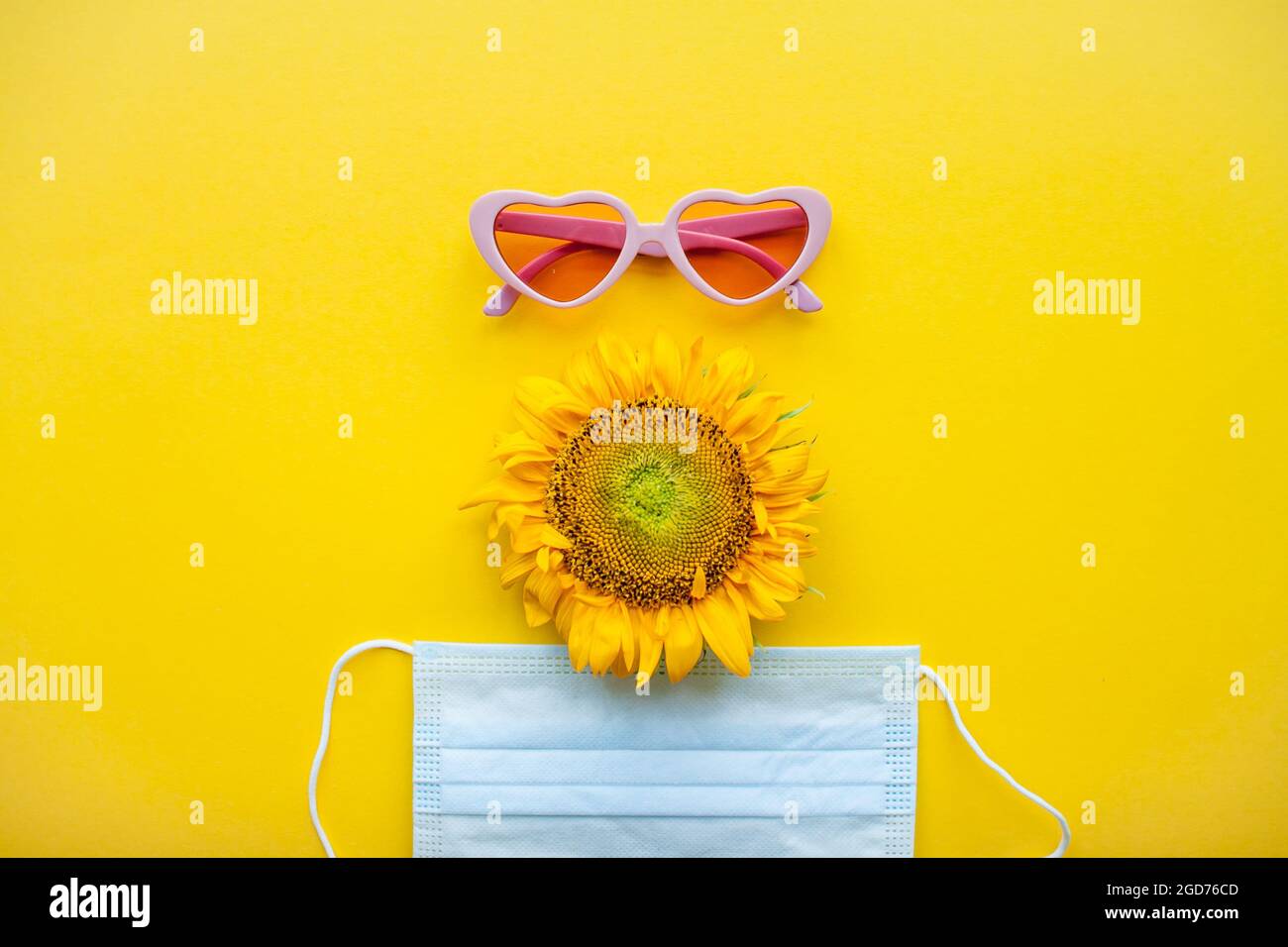 Funny face made of pink heart-shaped sunglasses, a protective medical mask and a sunflower on a bright yellow background Stock Photo