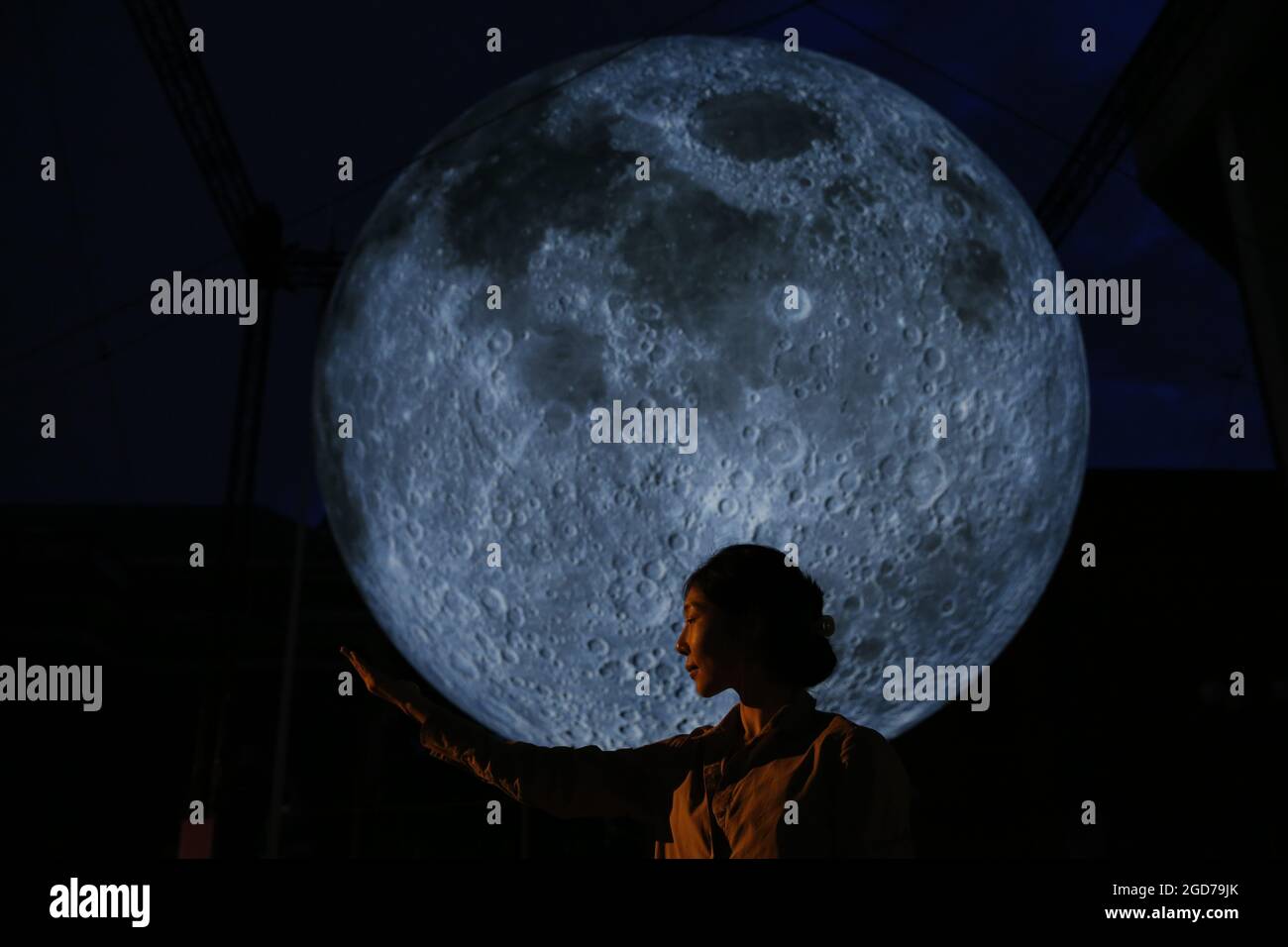A woman poses for a photo with Luke Jerram's 'Museum of the Moon', a light and sound installation that includes a replica of the moon seven metres in diameter, hangs in Kensington Town Square in London as part of the Kensington + Chelsea Festival. Picture date: Wednesday August 11, 2021. Stock Photo