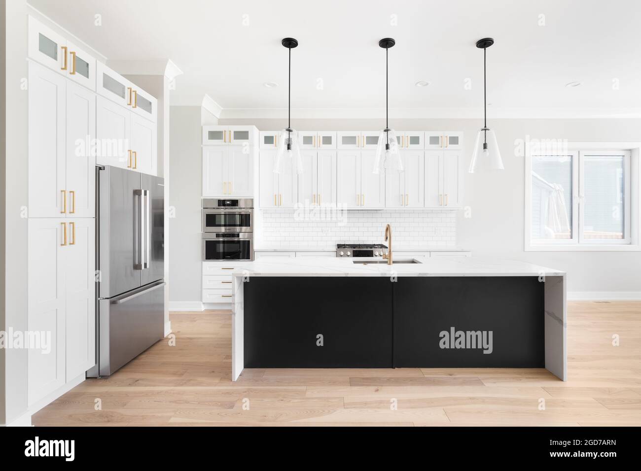 A luxury white kitchen with black pendant lights hanging above a waterfall granite island, stainless steel Bosch appliances, and gold hardware. Stock Photo