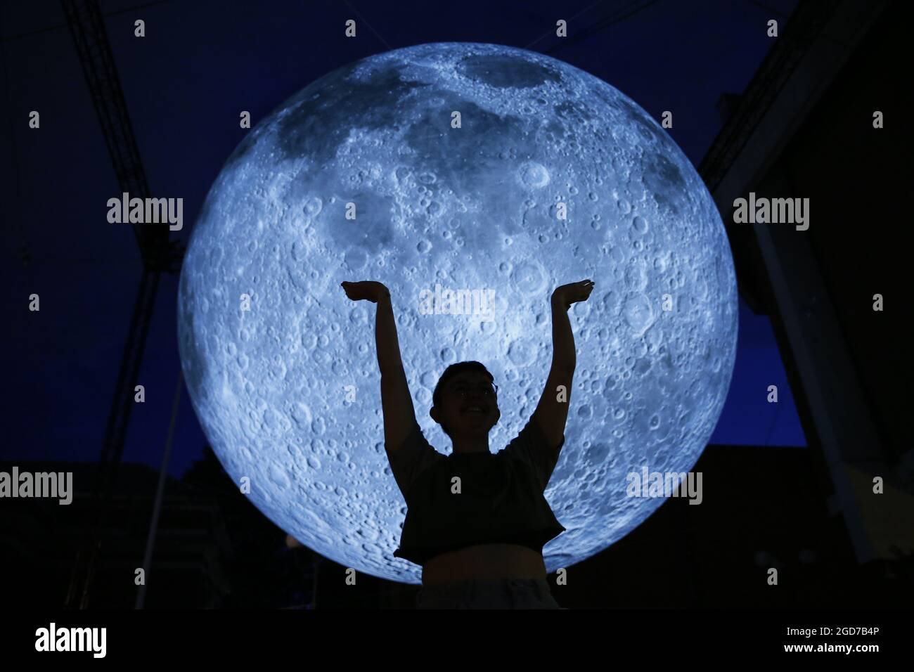 A person poses for a photo with Luke Jerram's 'Museum of the Moon', a light and sound installation that includes a replica of the moon seven metres in diameter, hangs in Kensington Town Square in London as part of the Kensington + Chelsea Festival. Picture date: Wednesday August 11, 2021. Stock Photo
