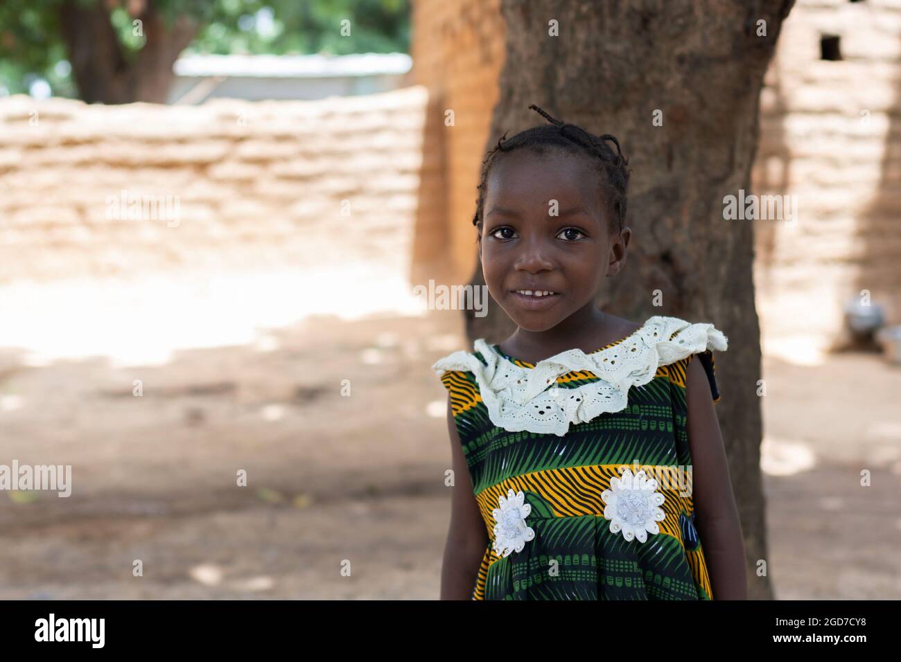 Adorable little black African girl standing before a tree in the courtyard of her home in a remote African village; poverty concept Stock Photo