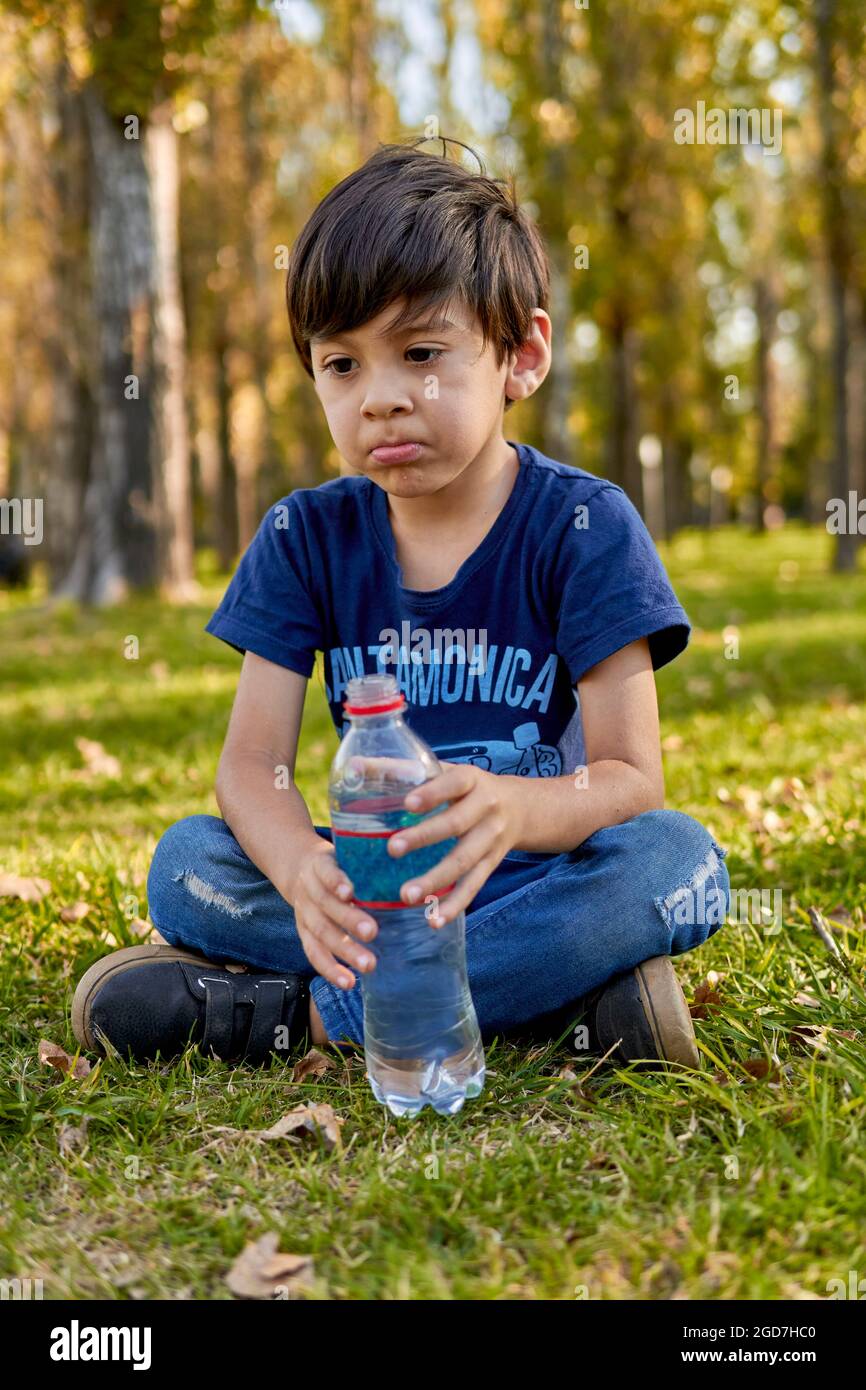 Brunette kid pensively holding a small bottle of water sitting in the middle of the forest at autumn. vertical, blurred background Stock Photo
