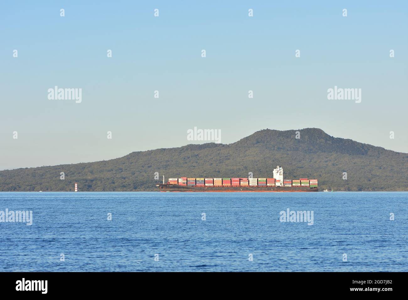 Container ship with rusty hull loaded with colourful shipping containers moving along island coast. Stock Photo