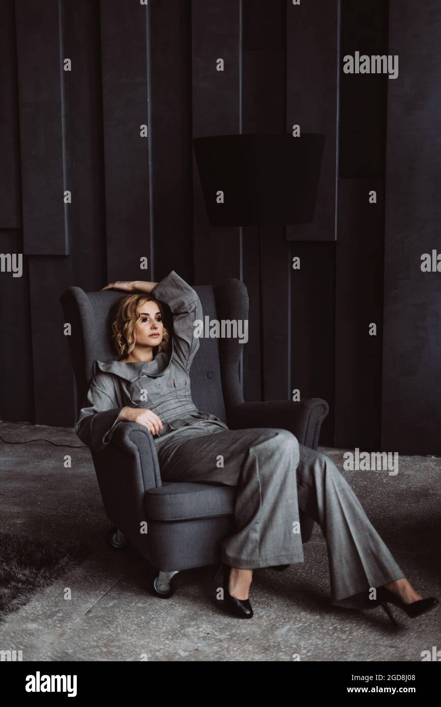 Beautiful young businesswoman in a stylish gray suit sitting on a chair in an office with a modern interior, looking at the camera. Portrait of a char Stock Photo