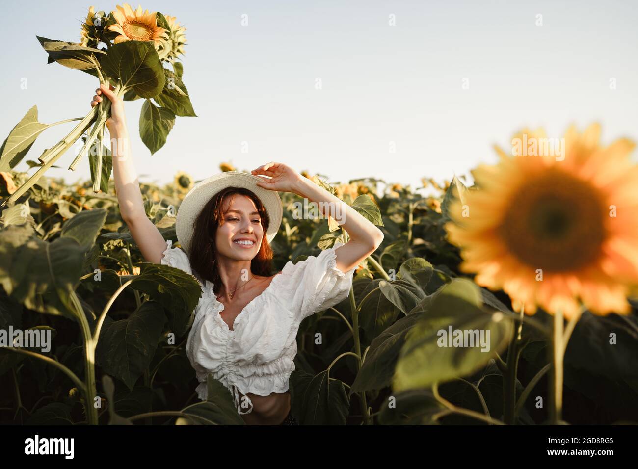 Smiling woman wearing a hat standing in a field of sunflowers in sunset ...