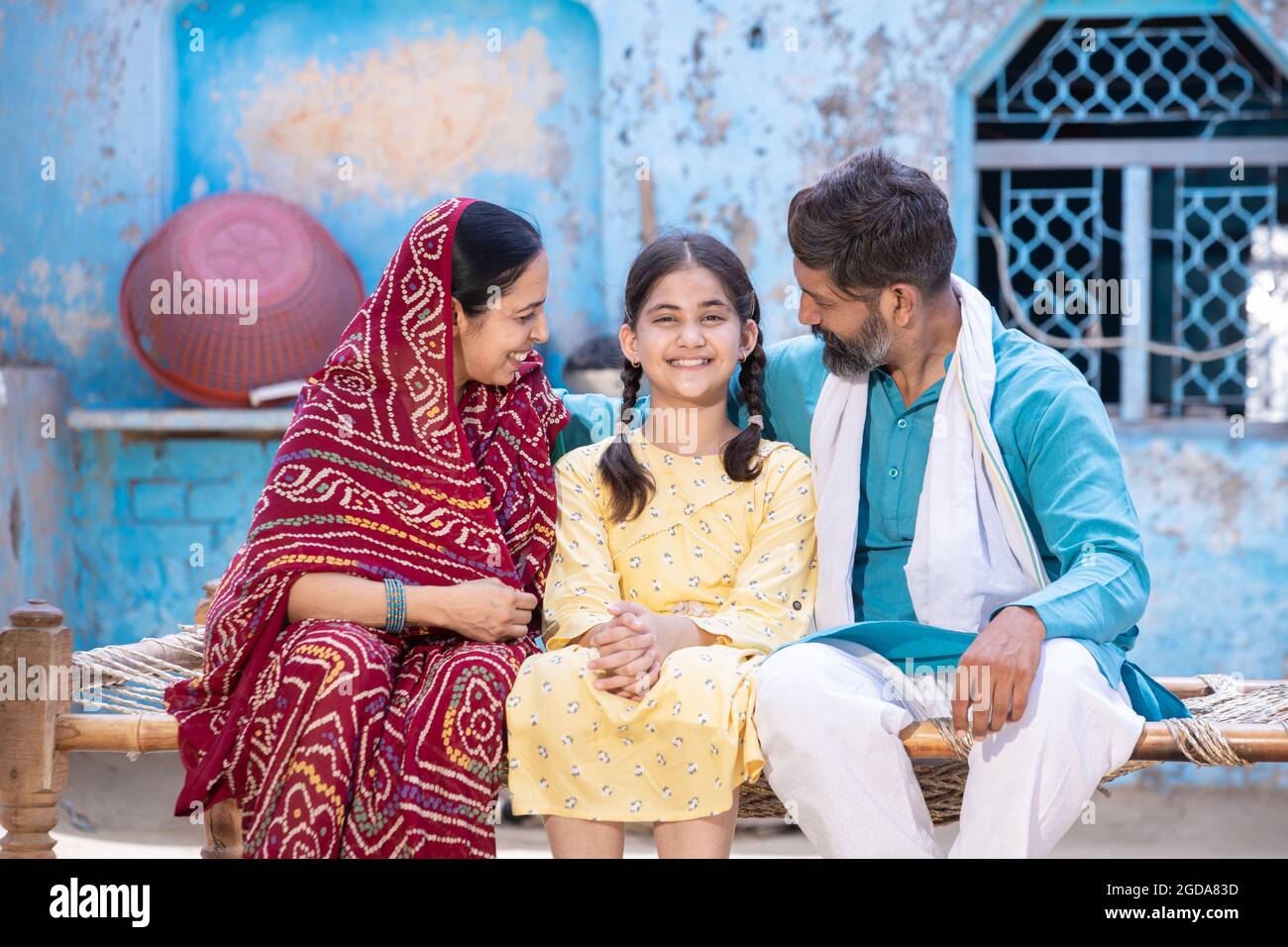 Happy rural indian family smiling while sitting on traditional bed at village home, India Village parent with Cute adorable little daughter smiling an Stock Photo