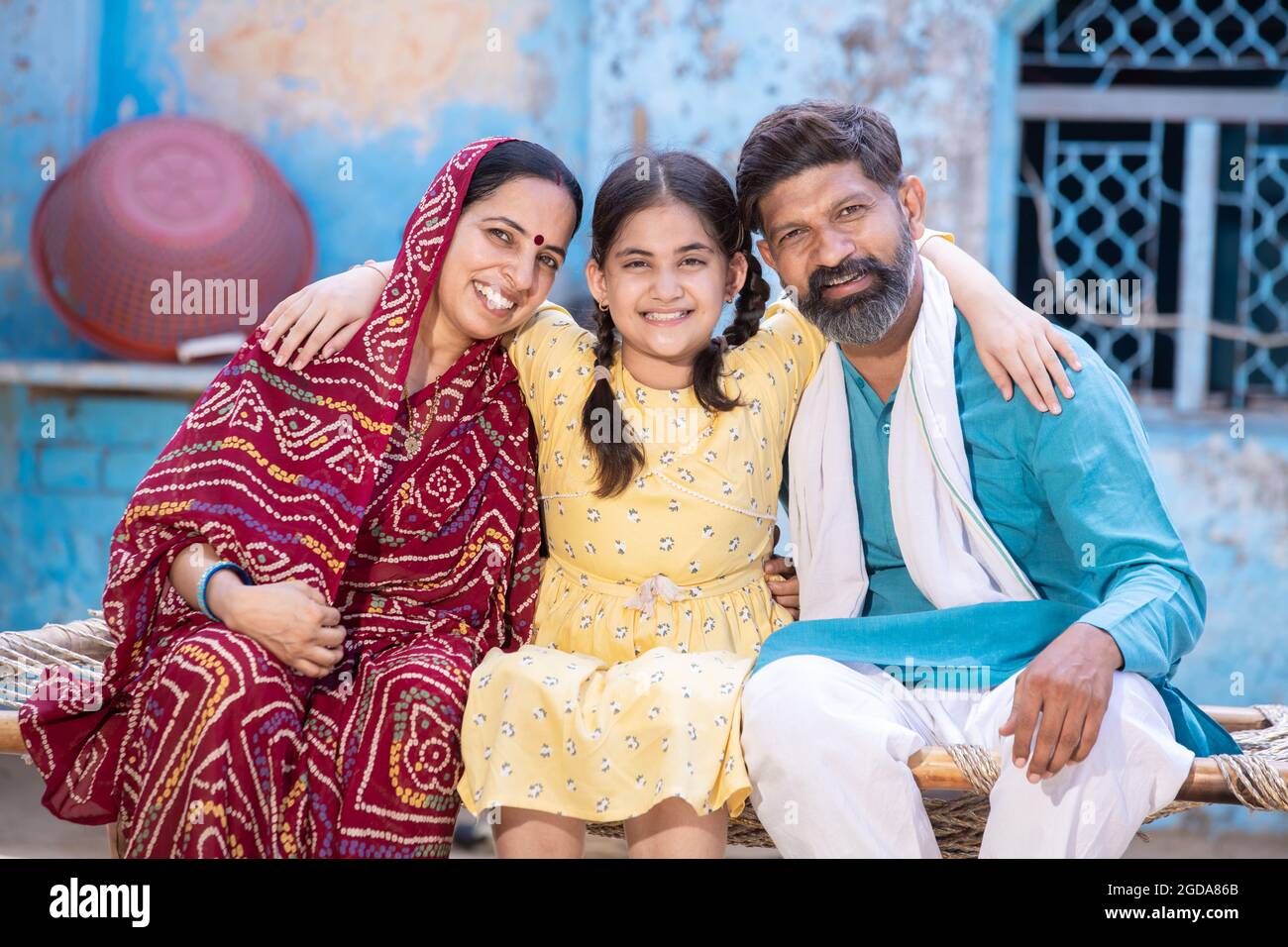 Portrait of happy rural indian family in looking at camera while sitting on traditional bed at village home, Cute adorable little daughter hug her fat Stock Photo