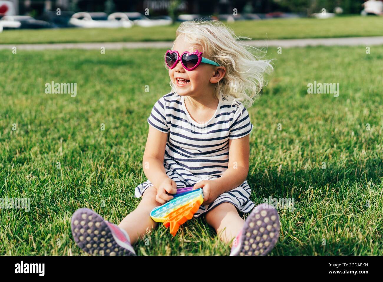 Cheerful emotional laughing little blonde girl with hair developing from the wind sits on the lawn. Stock Photo