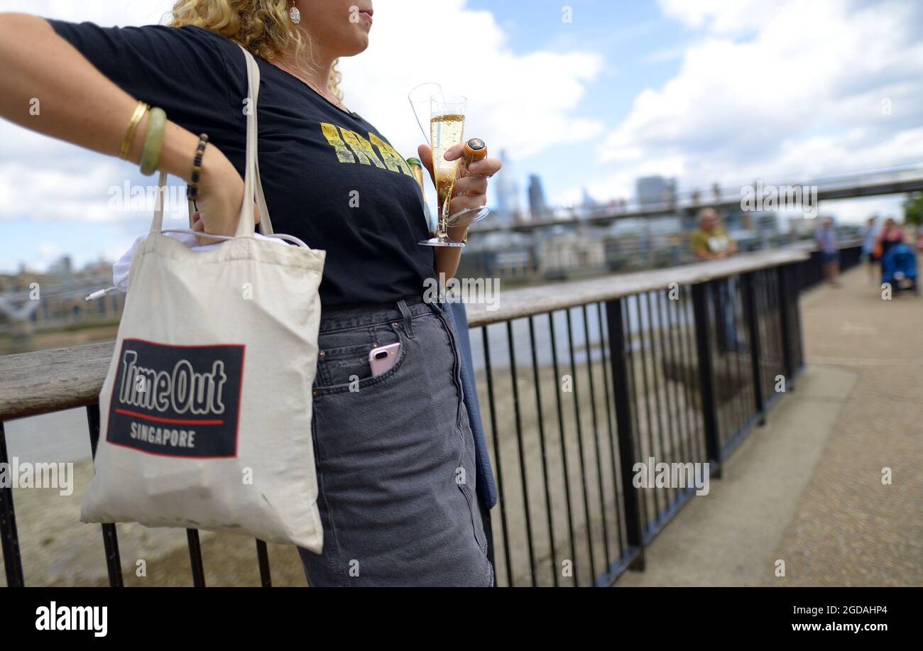 London, England, UK. Woman drinking champagne form a plastic champagne flute outdoors on the South Bank with a Time Out bag Stock Photo