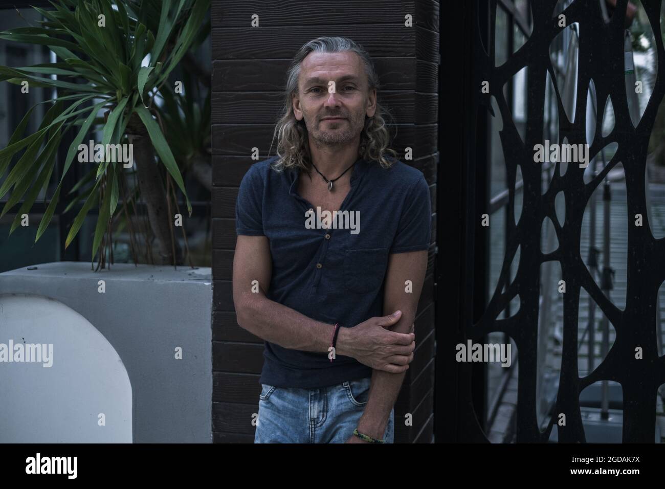 Tanned mature handsome man in t-shirt and jeans standing on the street. Long grey hair. Portrait. Grunge style. Looking at camera with copy space. Stock Photo