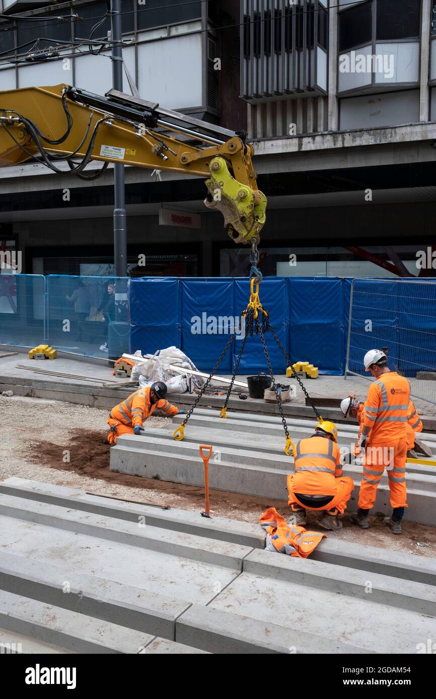 Team of workmen wearing orange high-viz overalls on a construction site work at laying the new track foundations for the update to the Midland Metro tram public transport system in the city centre along Corporation Street on 3rd August 2021 in Birmingham, United Kingdom. The original tracks are being pulled up and relaid, while a new line is also under construction and due to open later in the year. The Midland Metro is a light-rail tram line in the county of West Midlands, England, operating between the cities of Birmingham and Wolverhampton via the towns of West Bromwich and Wednesbury. The Stock Photo