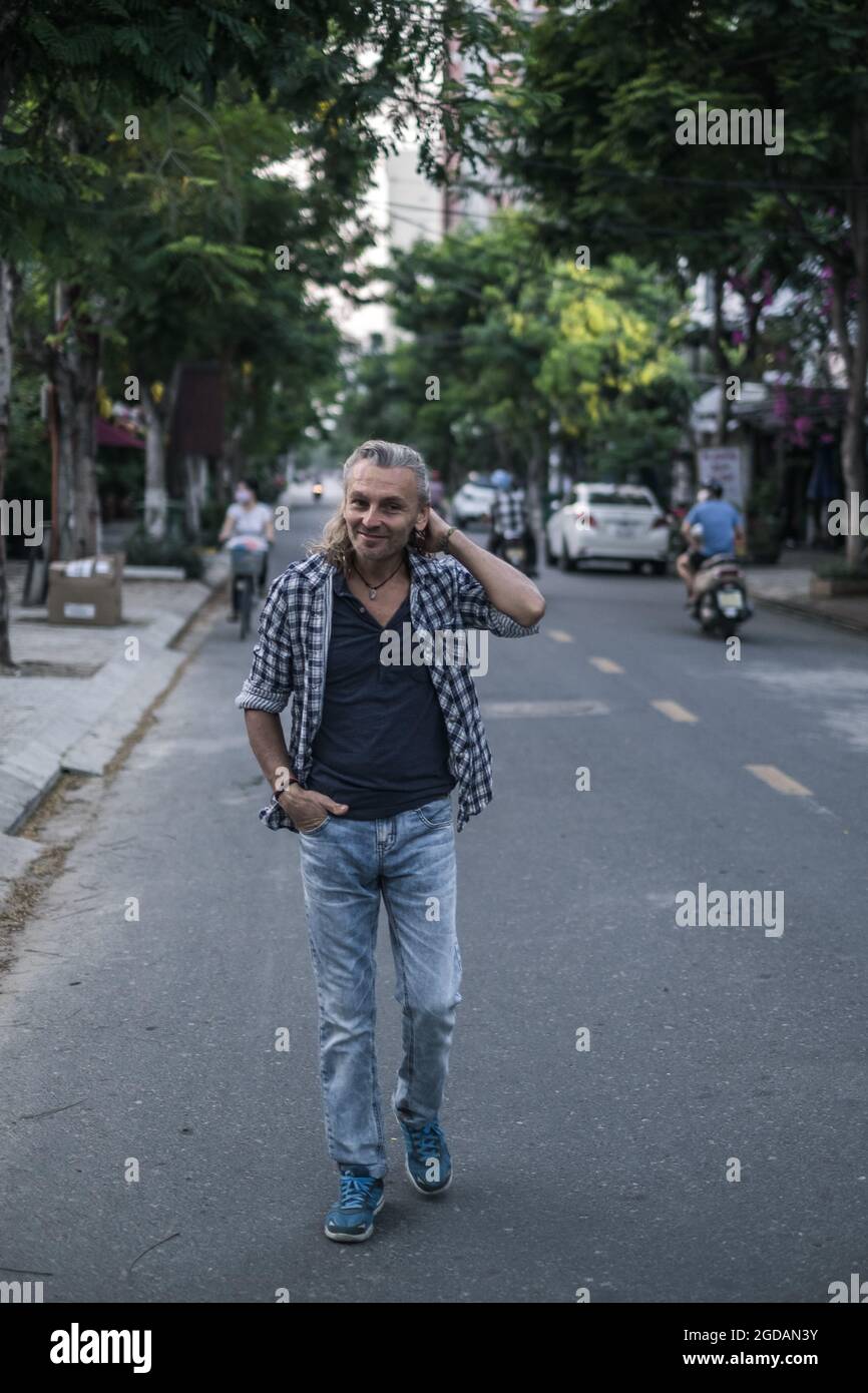 Elderly man in t-shirt, jeans walking on the street in town and holding his hair. Long grey hair. High quality photo Stock Photo