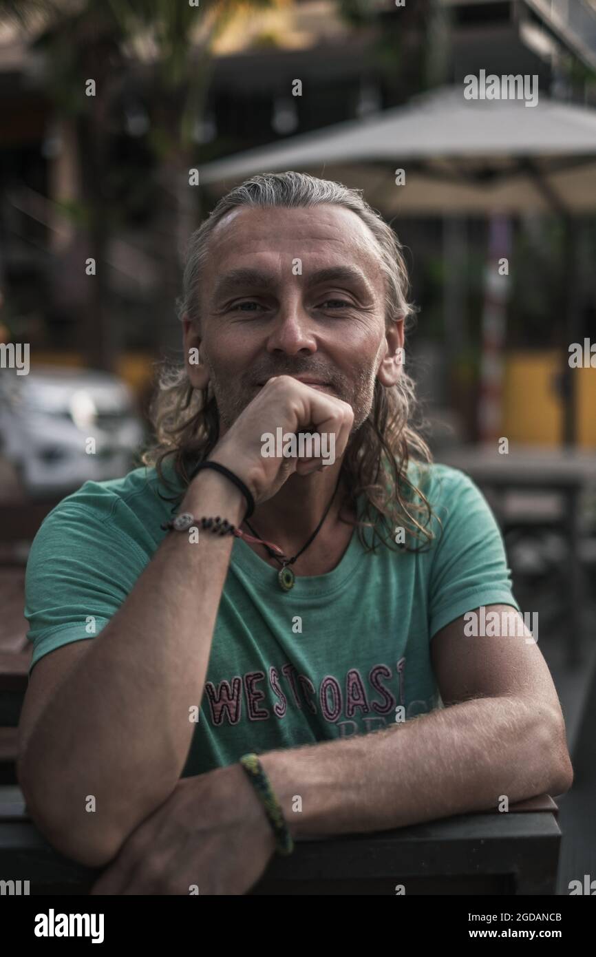 Close-up portrait of mature handsome man sitting on the summer veranda of the restaurant, holding his hand to his chin. Long grey hair. High quality Stock Photo
