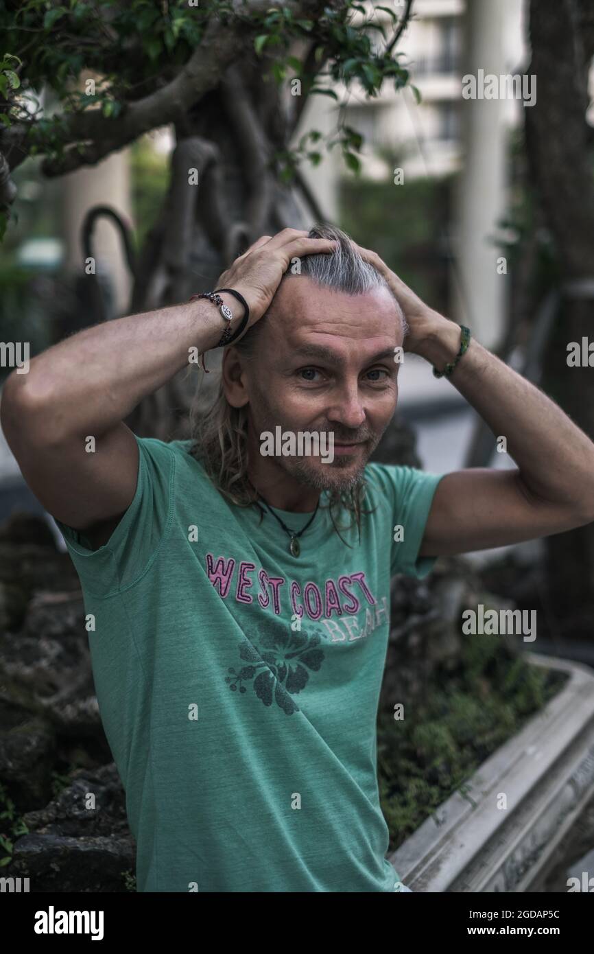 Smiling elderly man with long grey hair wearing green t-shirt sitting on the street in town and holding his hair with two hands. Portrait. copy space. Stock Photo