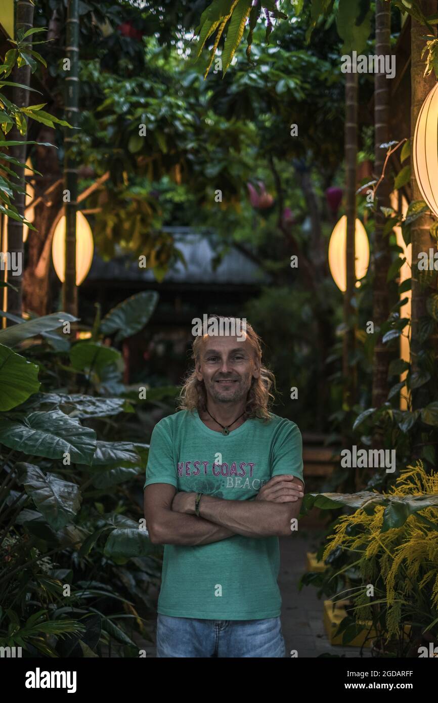 Portrait of mature man in t-shirt, jeans standing on the green street. Long grey hair. Chinese lanterns. Evening. Trees on the background. Stock Photo