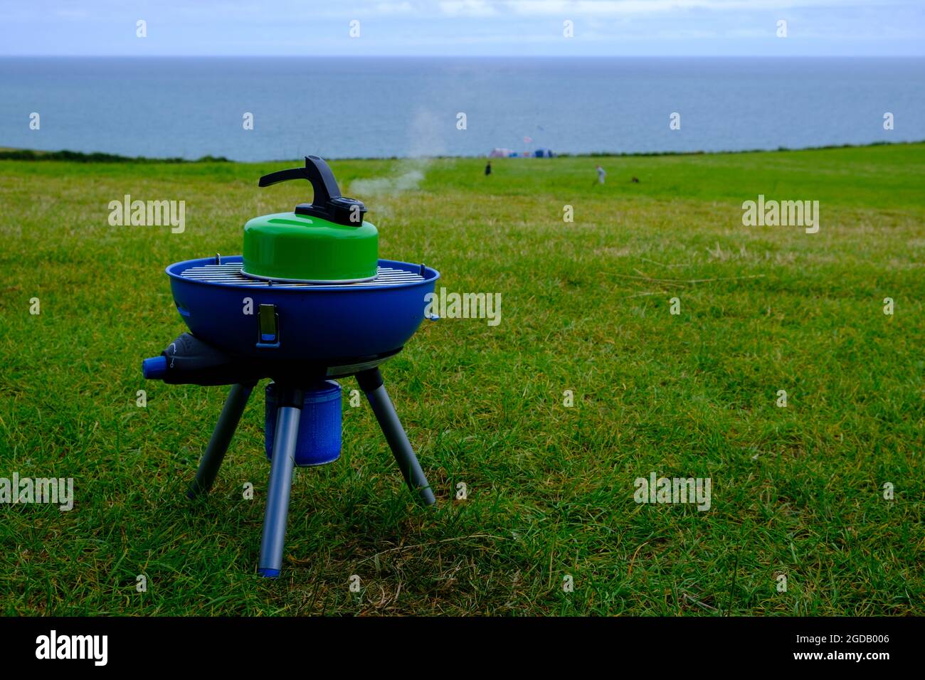Blue Campingaz camping stove with a green boiling kettle with steam coming out of the spout and the sea in the background.2021 Stock Photo