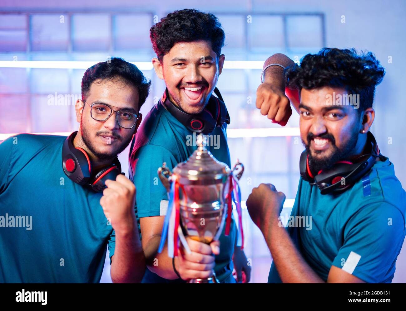 Team of Excited Cheerful gamers with headphones celebrating by dancing and holding winning trophy at esports gaming tournament stage Stock Photo