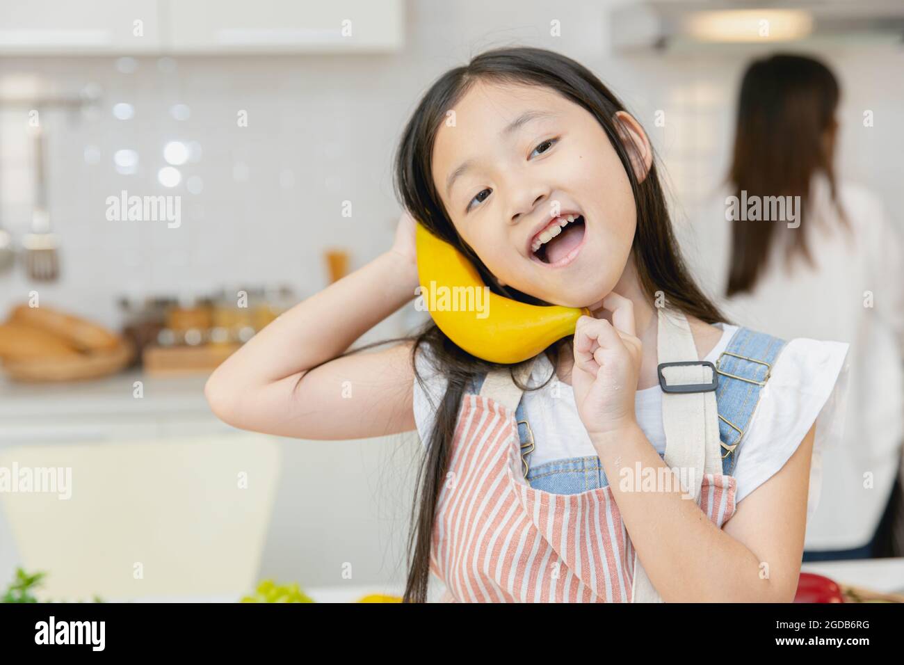 Cute girl child enjoy funny candid moment playing with banana at home kitchen. Stock Photo