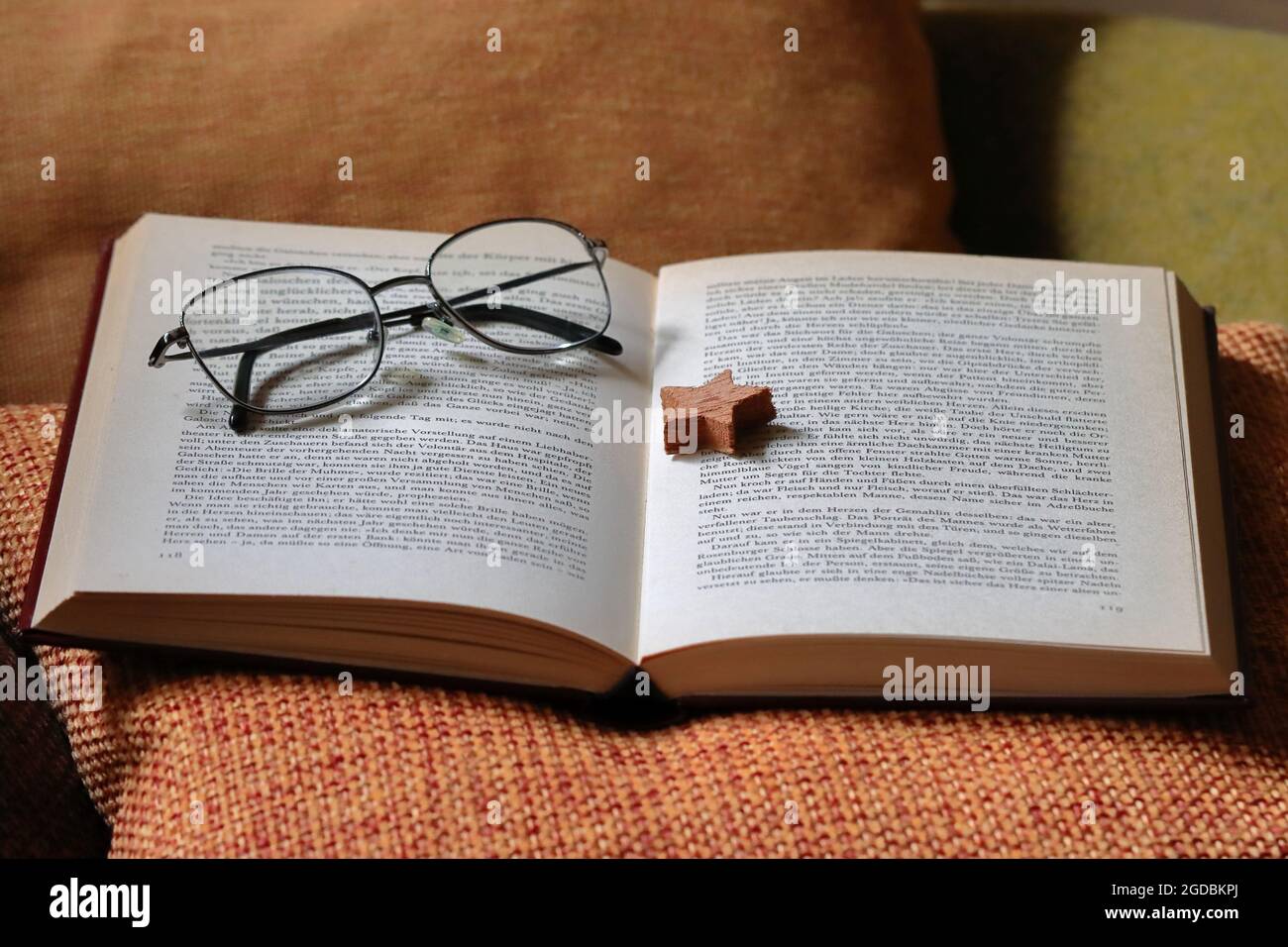 ein aufgeschlagenes Buch liegt zusammen mit einem Holzstern als Lesezeichen und einer Brille auf einem orangenen Kissen Stock Photo