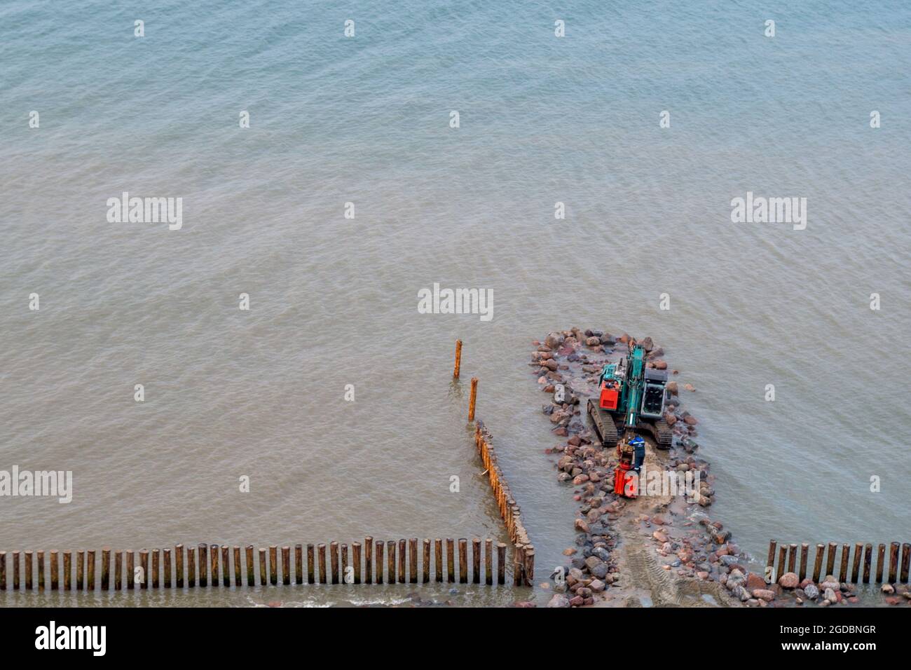 Blue excavator with red working part, standing on base of large stones is involved in formation of wooden breakwater from piles protect shore on backg Stock Photo