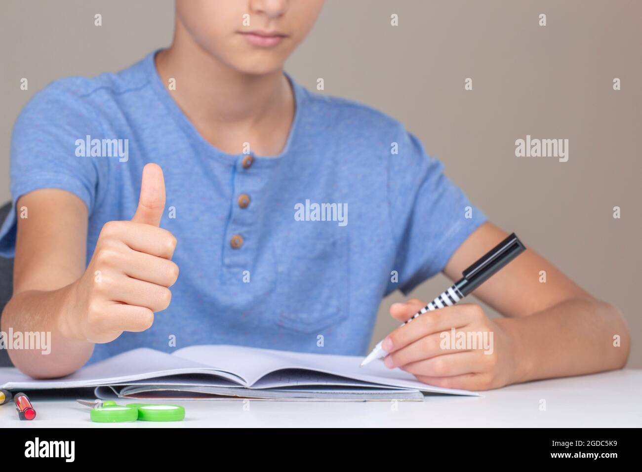 Boy hand holding pen in left hand and writing in a notebook, doing homework. Thumb up hand gesture. Left Handers Day Stock Photo