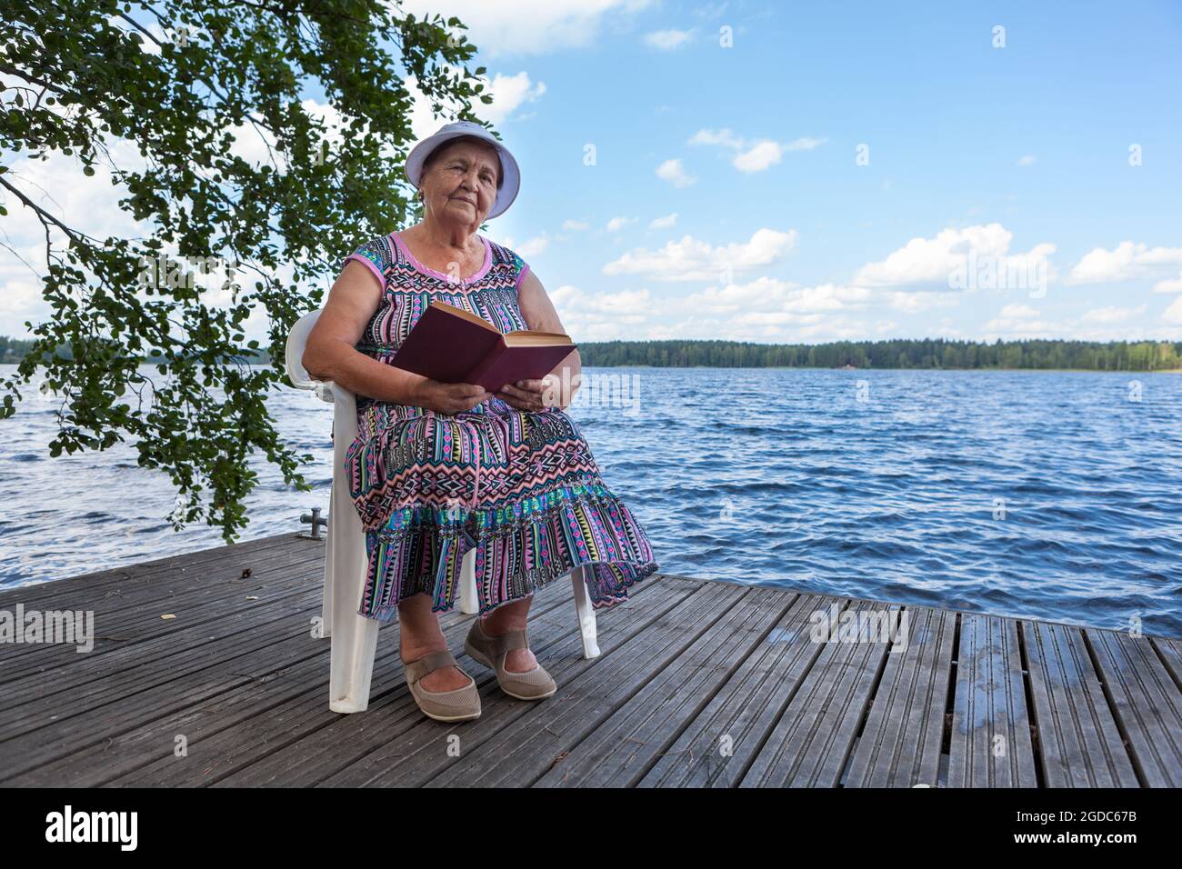 Old lady reading book sitting at the plastic chair in forest lake shore, looking at camera Stock Photo