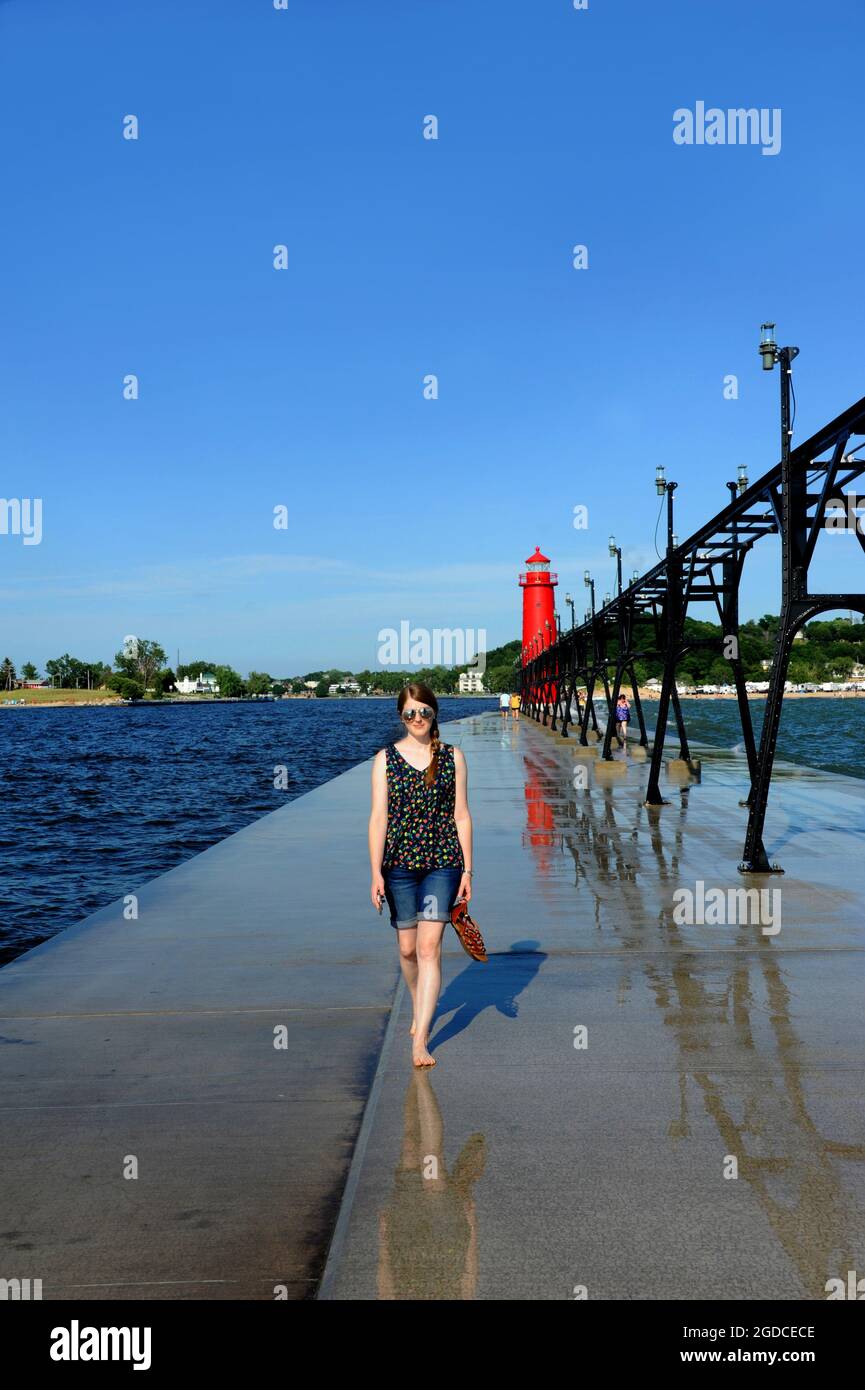 Attractive young woman walks barefoot along the pier.  The red Grand Haven Lighthouse stands in the background. Stock Photo