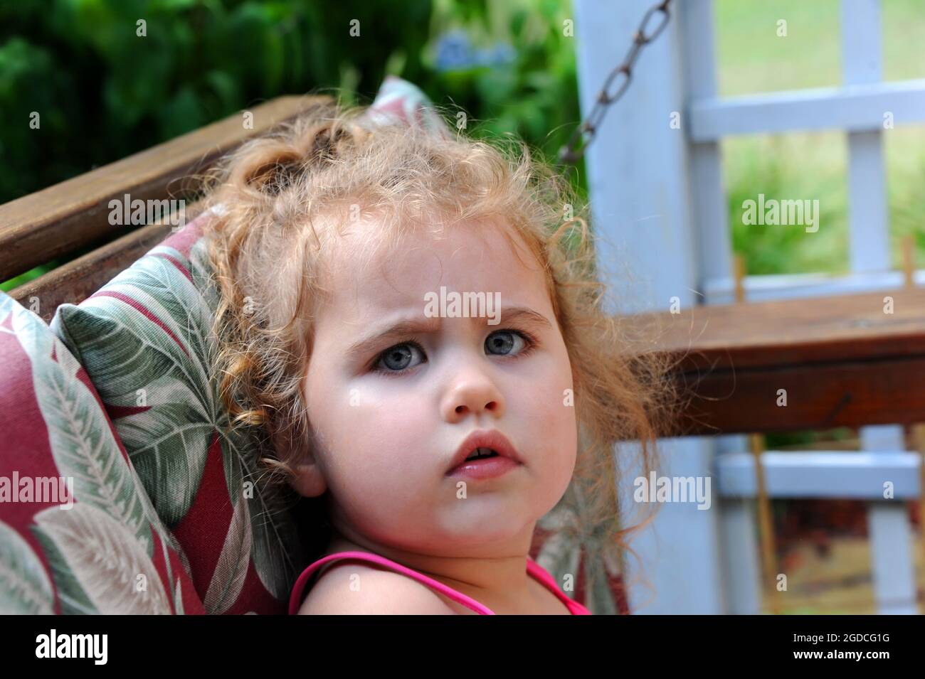 Frown lines perch on a sweet faced little girl.  she is crabby and grouchy as she sits on her front porch swing.  She has curly hair and a ponytail. Stock Photo