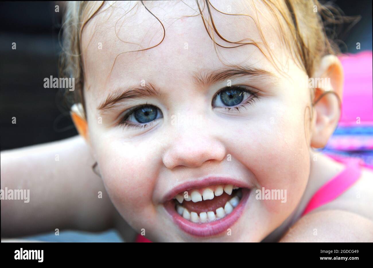 Closeup shows a little girl's wet face.  She has tendrils of hair clining to her forehead.  Her mouth is open in a wide smile. Stock Photo