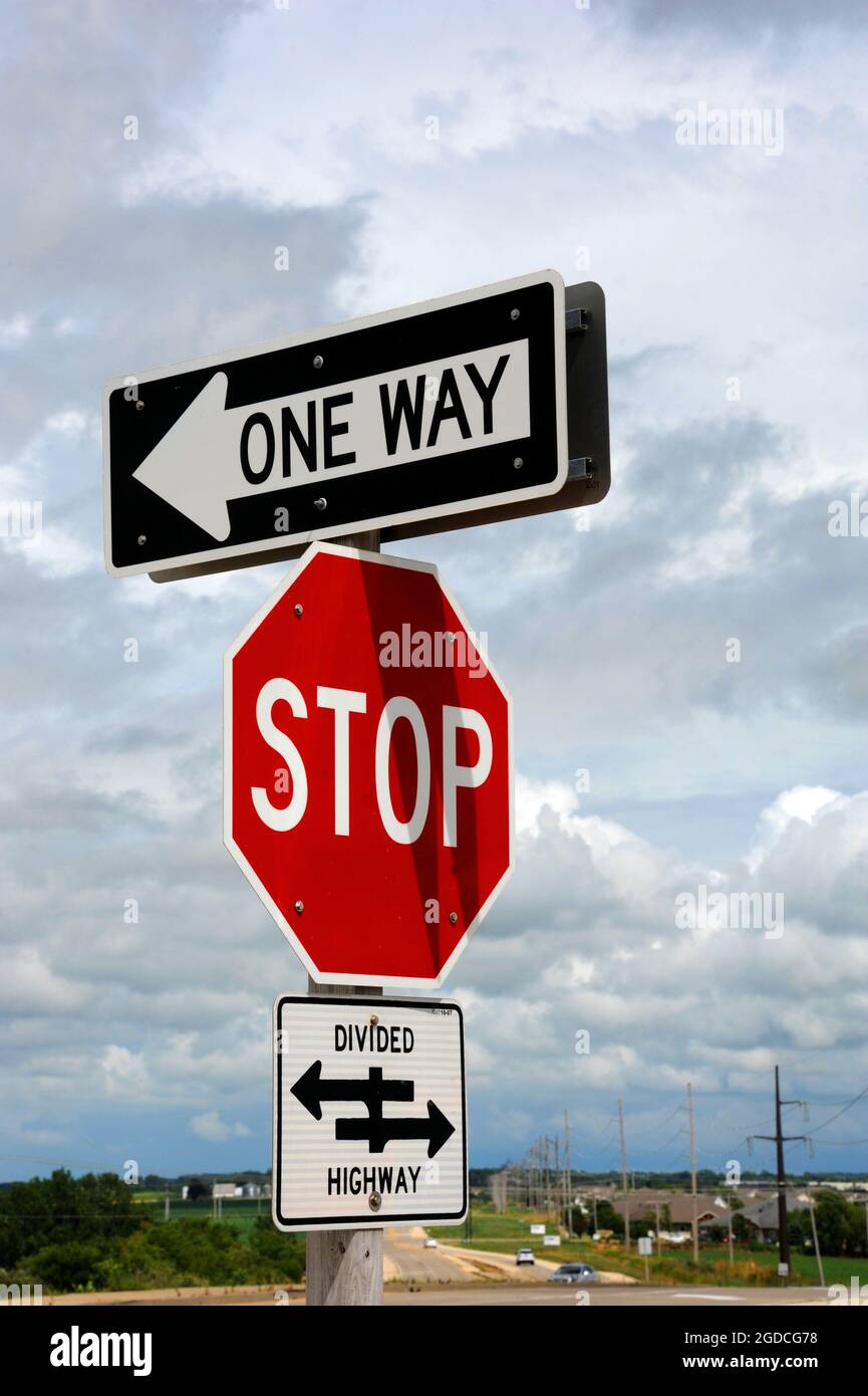 Three road signs sit one on top of the other.  One says 'one way', another Stop, and the bottom sign indicates a divided highway. Stock Photo