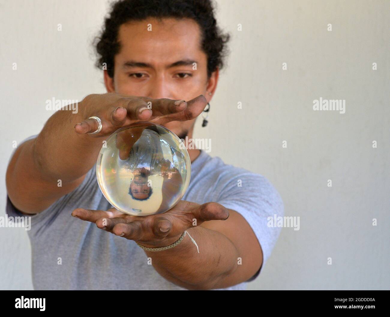 Handsome young man with dirty fingernails holds in his hands a crystal ball with his own vertically flipped mirror image and looks at the orb. Stock Photo