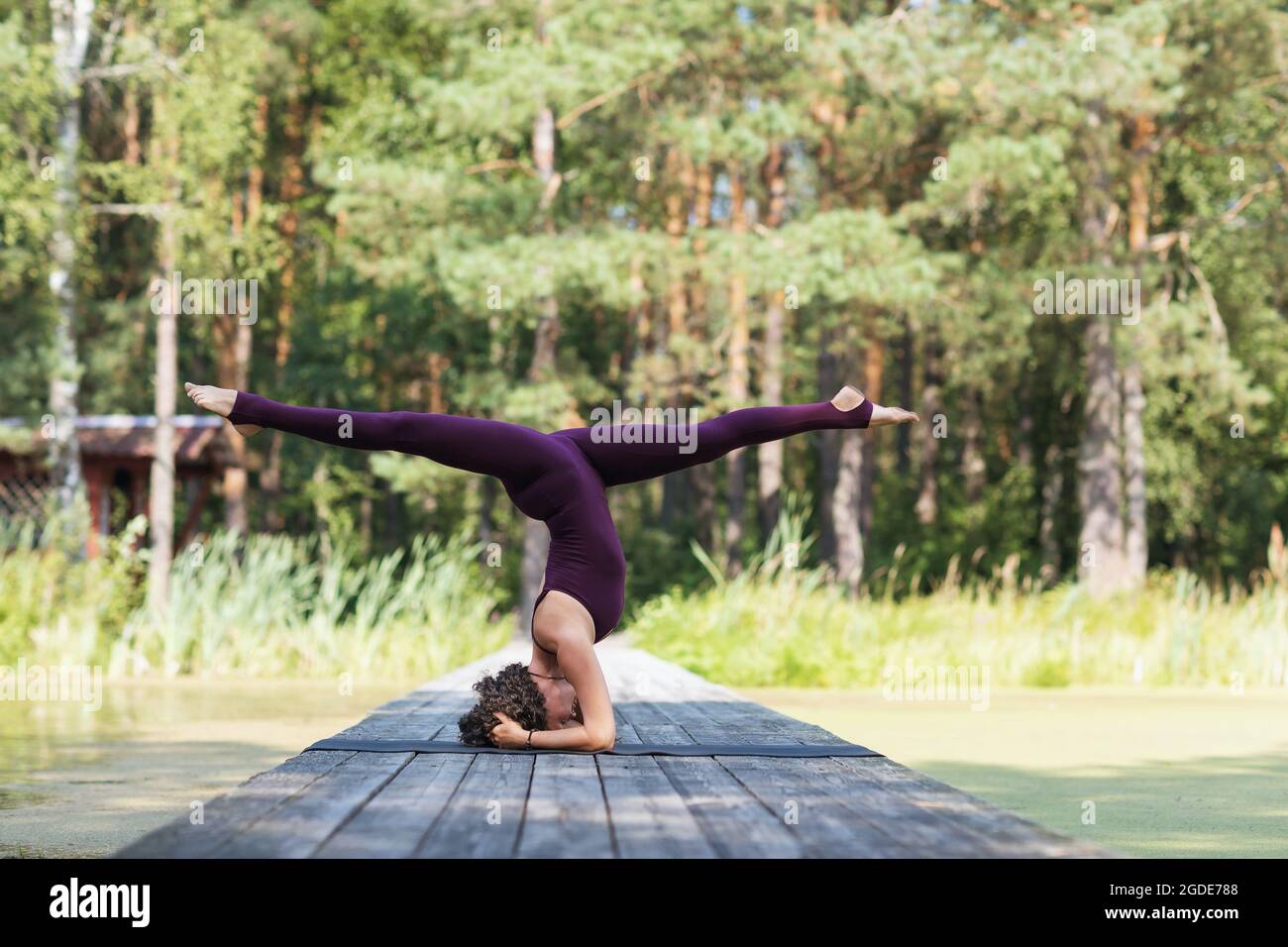A woman in sportswear, practicing yoga in the park, on a mat performs a shirshasana exercise with a transverse twine, headstand Stock Photo