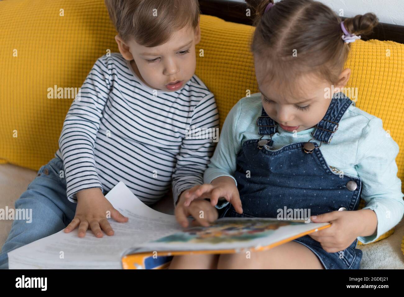 Two preshool, toddler children read large interesting book of fairy tales on yellow bed. Siblings little twins reader have fun, happy kid on Stock Photo