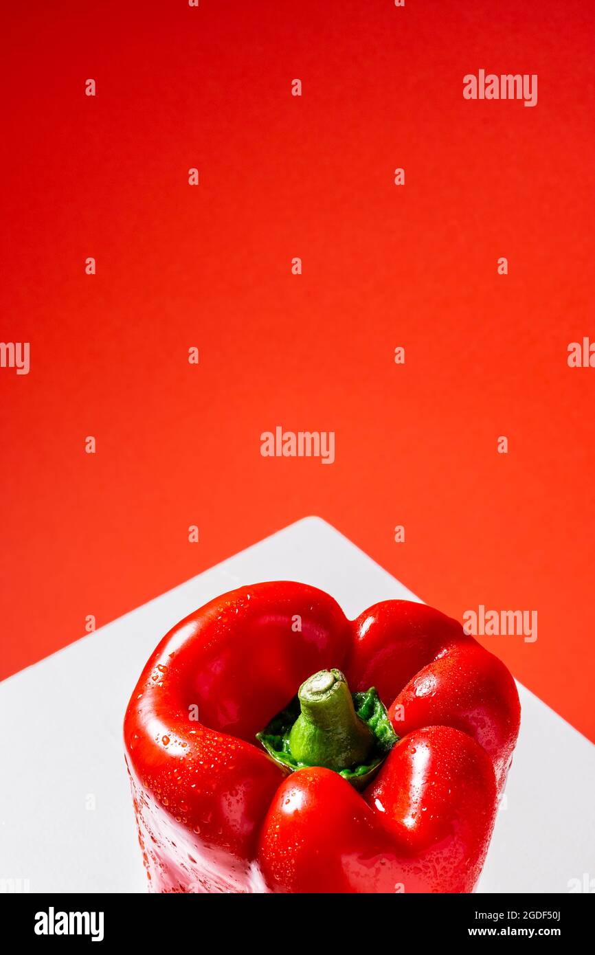 Photo of a wet red bell pepper on a white table and red background from a top point of view.The photograph is taken in vertical format and has space t Stock Photo