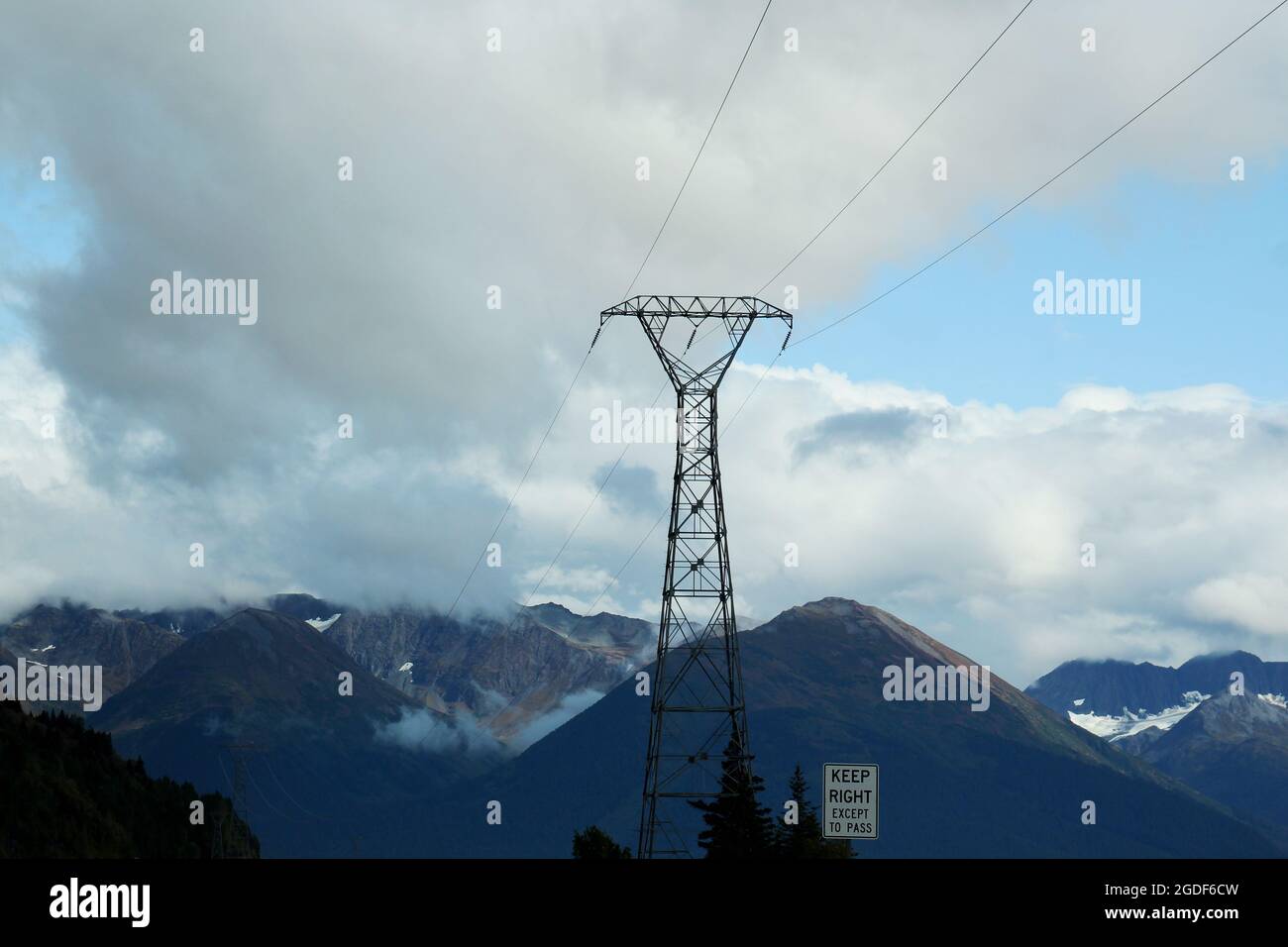 Ein großer Strommast mit Stromleitungen und einem Hinweisschild 'Keep right expect pass' in einer Berglandschaft an einem bewölkten Tag in Alaska, USA. Stock Photo
