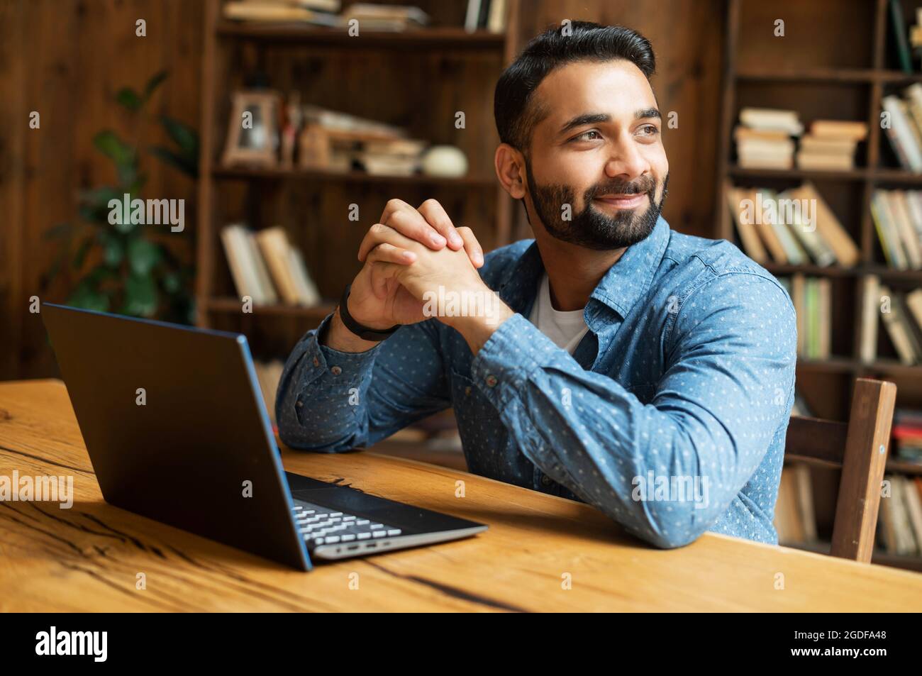 Happy and optimistic Indian man in casual wear using laptop in modern office, successful inspired arabic male entrepreneur looks away and smiles thoughtfully, planning next step Stock Photo