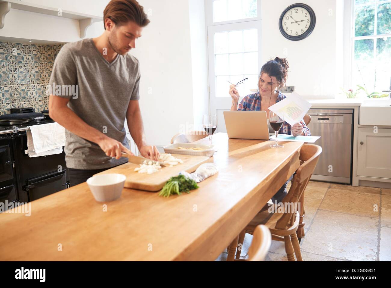 Man preparing vegetables at kitchen table, girlfriend using laptop Stock Photo