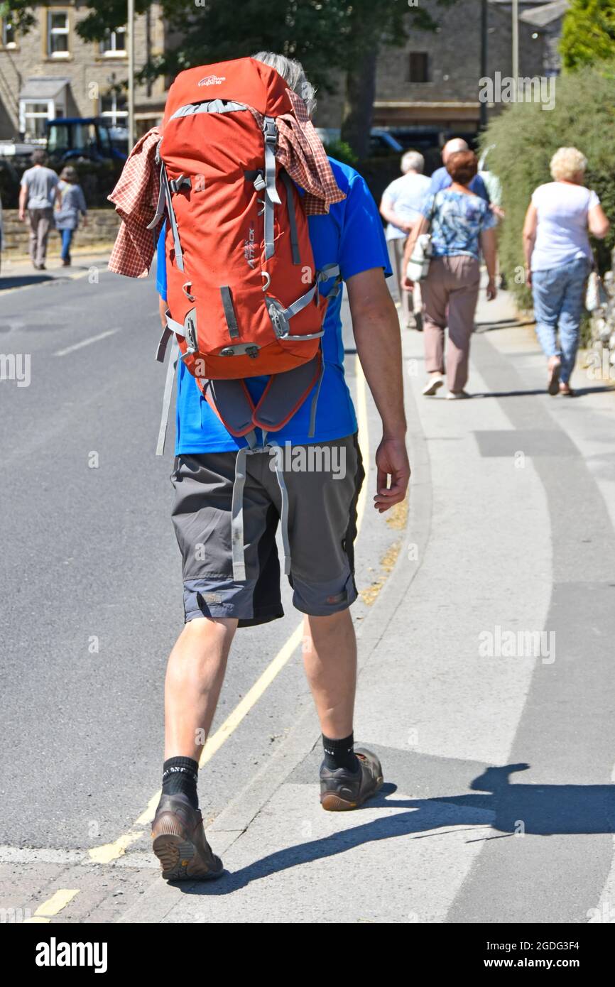 Close up back view Dales walker man carrying big backpack in shorts & walking boots on pavement in Grassington Wharfedale North Yorkshire England UK Stock Photo