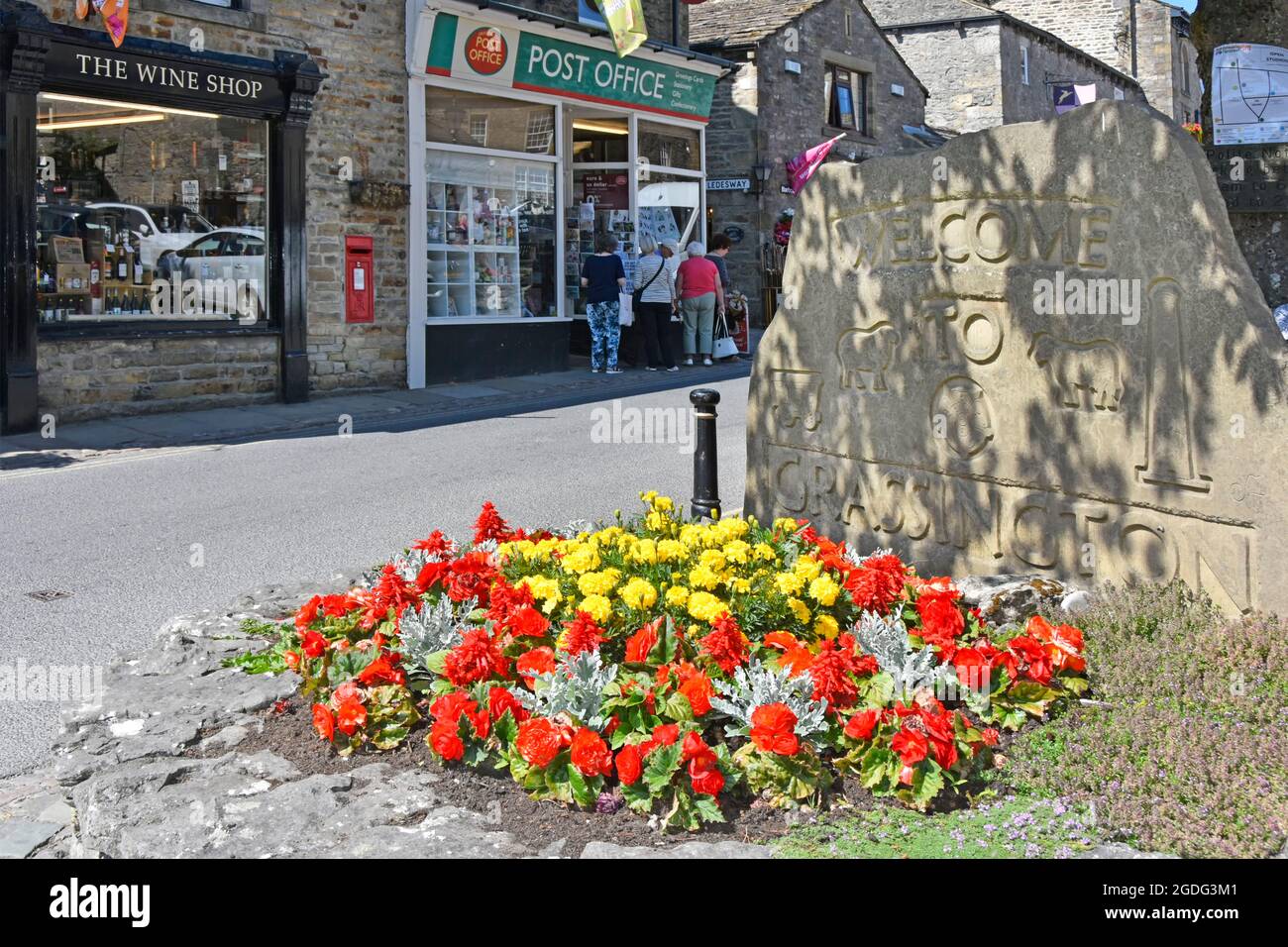 People & Post Office street scene beside colourful summer flower bed & stone welcome sign to Grassington village in Wharfedale North Yorkshire England Stock Photo
