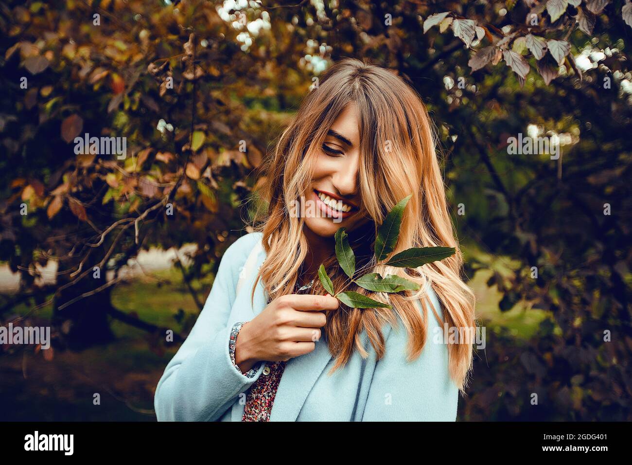Woman with long blond hair holding sprig of leaves in park, portrait Stock Photo