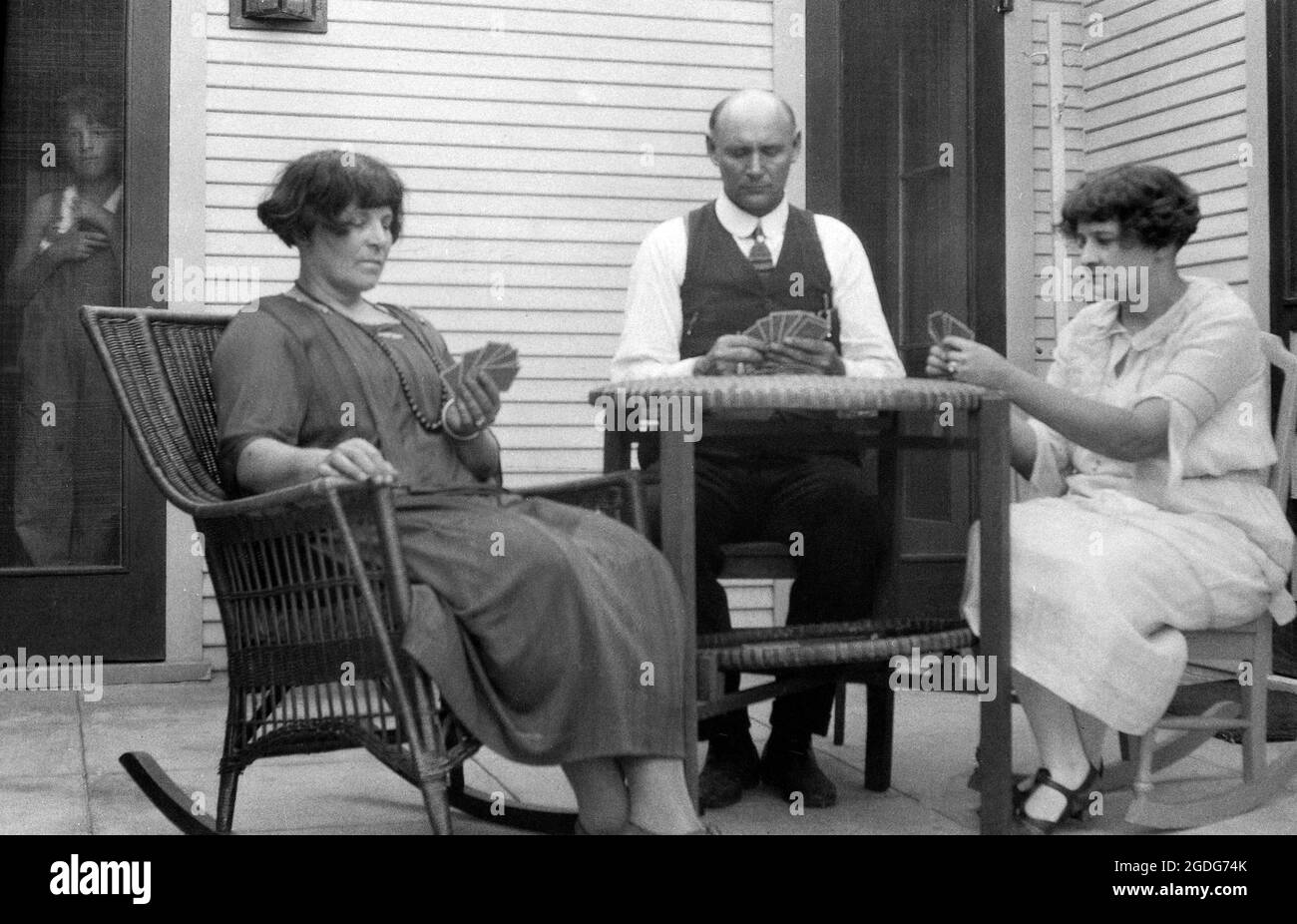 1930s, historical, a man and two ladies formally dressed in the clothes of the era, sitting outside at a table on a veranda playing cards, watched by a young boy at the doorway inside the house, USA. Stock Photo