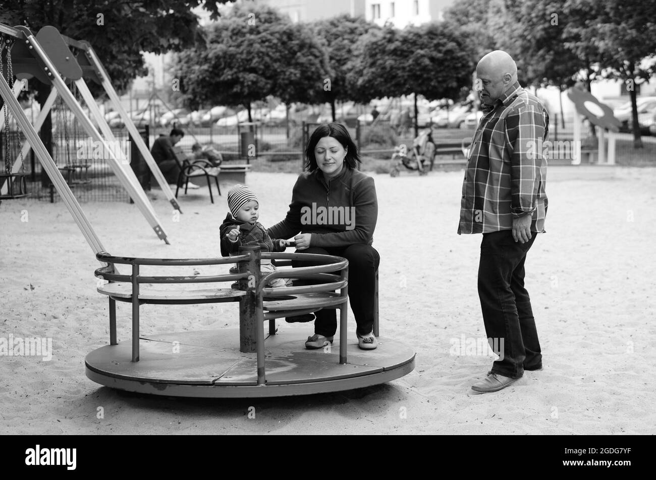 POZNAN, POLAND - May 16, 2015: Senior man standing next to woman with baby on roundabout at a play ground Stock Photo