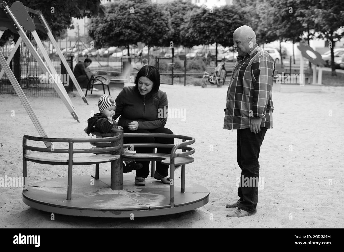 POZNAN, POLAND - May 16, 2015: Senior man standing next to woman with baby on roundabout at a play ground Stock Photo