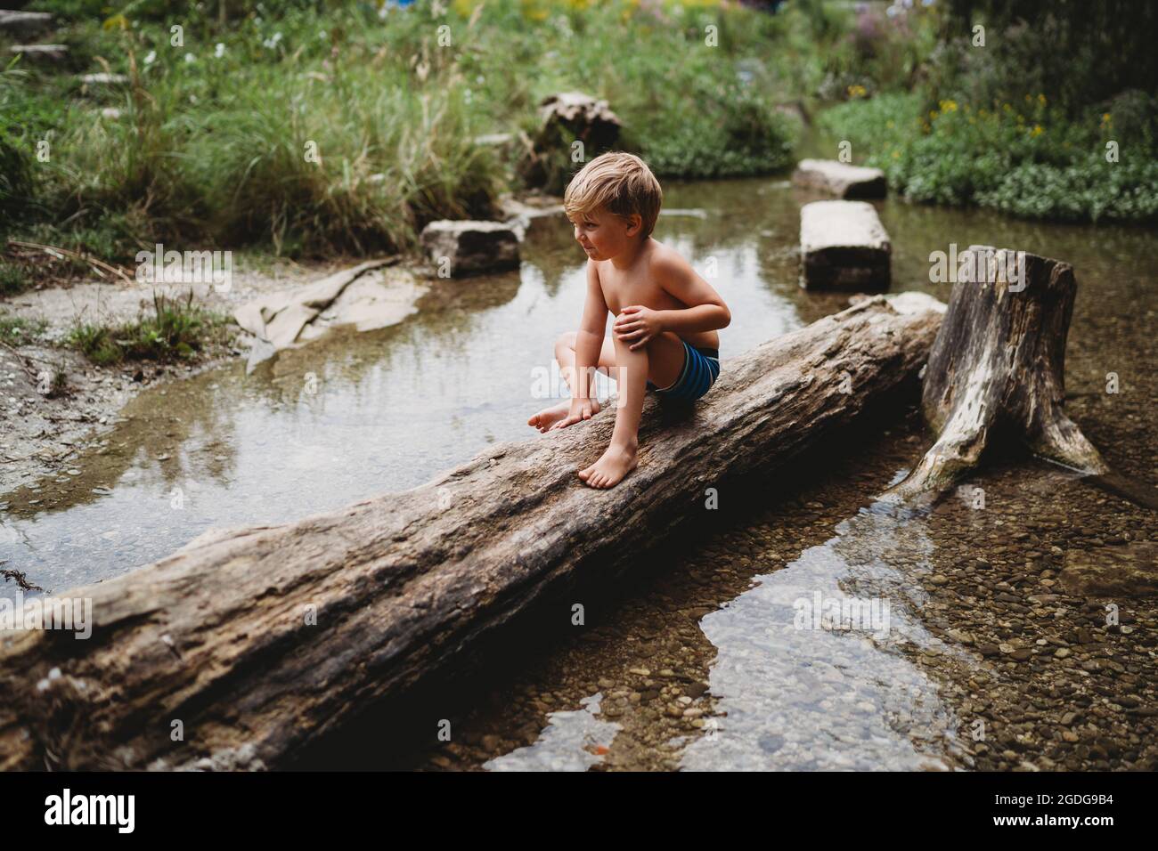 Young boy sitting on log in water in summer looking thoughtful Stock Photo