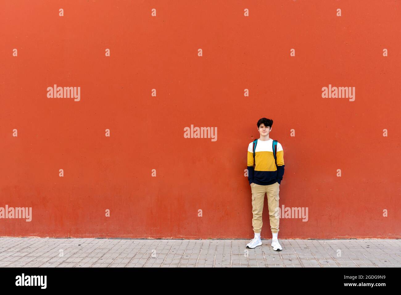 Young male with backpack standing on red wall while looking to camera Stock Photo